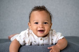 A happy baby wearing a white outfit lies on their tummy while smiling brightly at the camera.