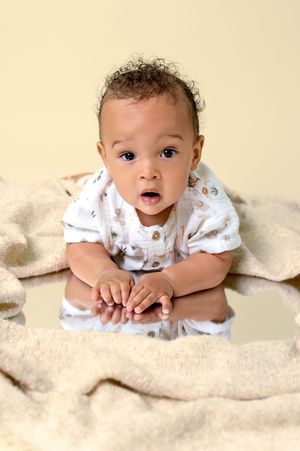 A baby in a white onesie lying on a beige blanket looking at the camera with bright eyes.