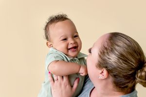 A joyful baby in a mint green shirt laughs while being lifted up against a beige background.