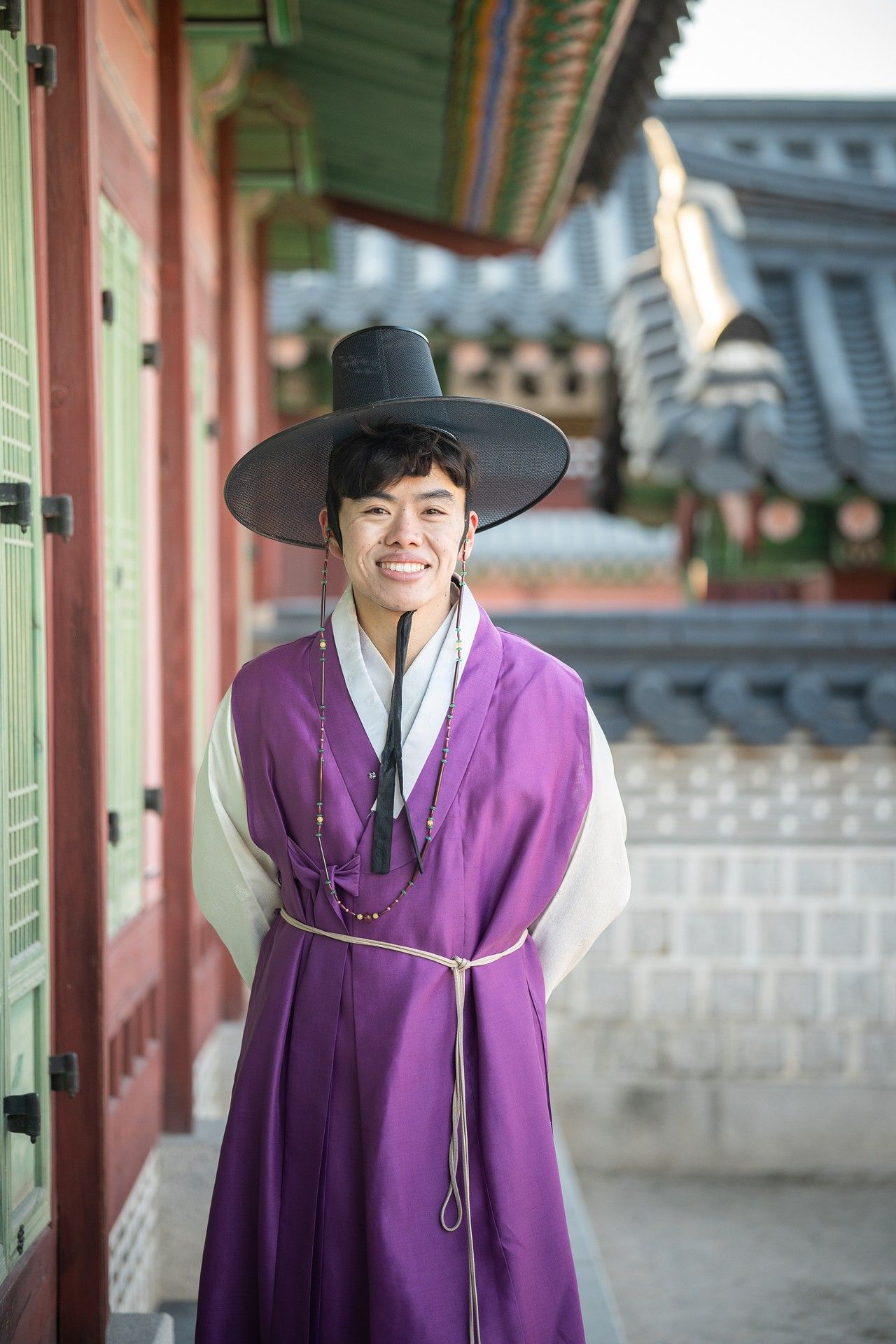 Person wearing traditional Korean hanbok in purple and black attire standing in front of historic palace architecture.
