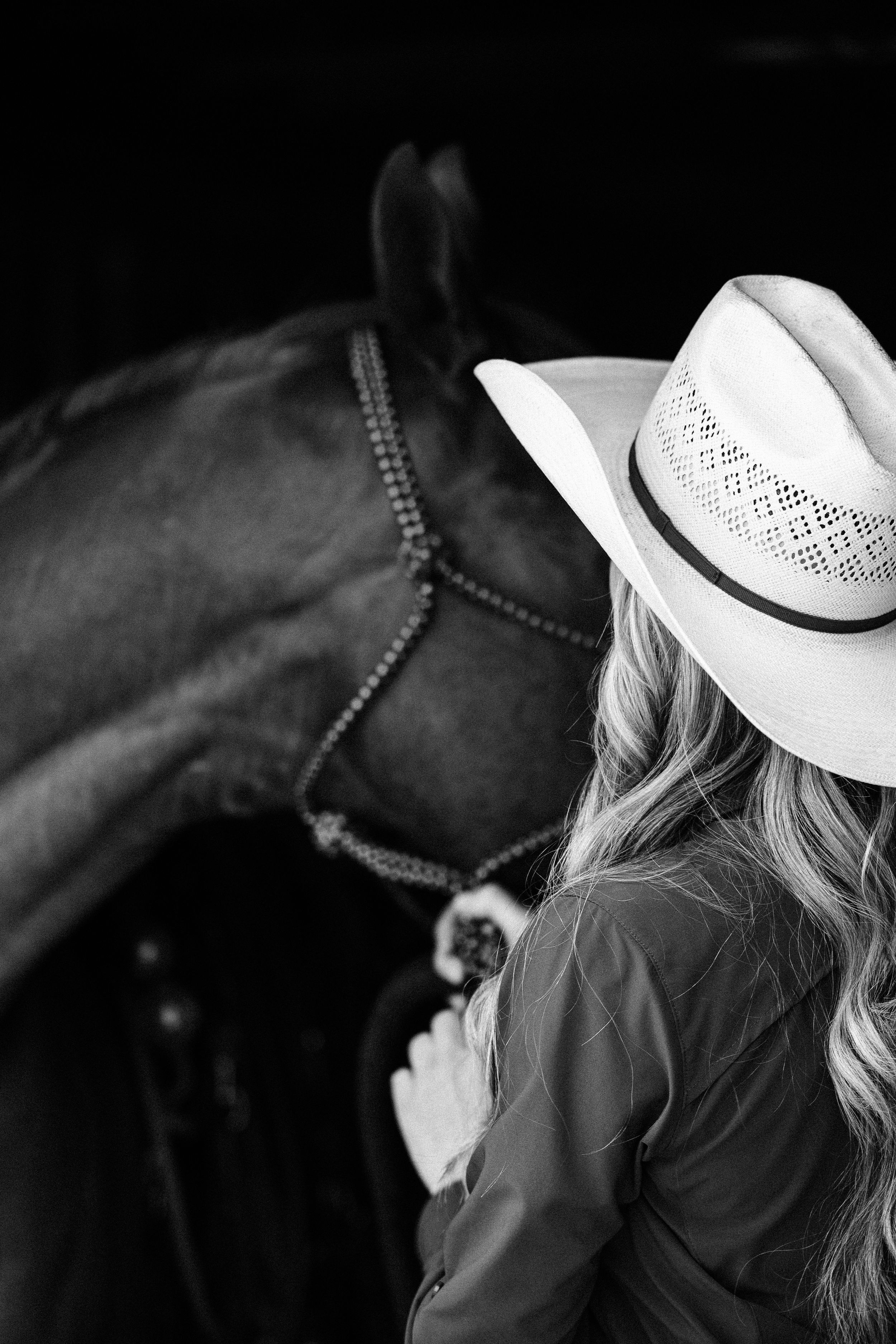 Black and white photo of blonde cowgirl in white hat standing beside dark horse.