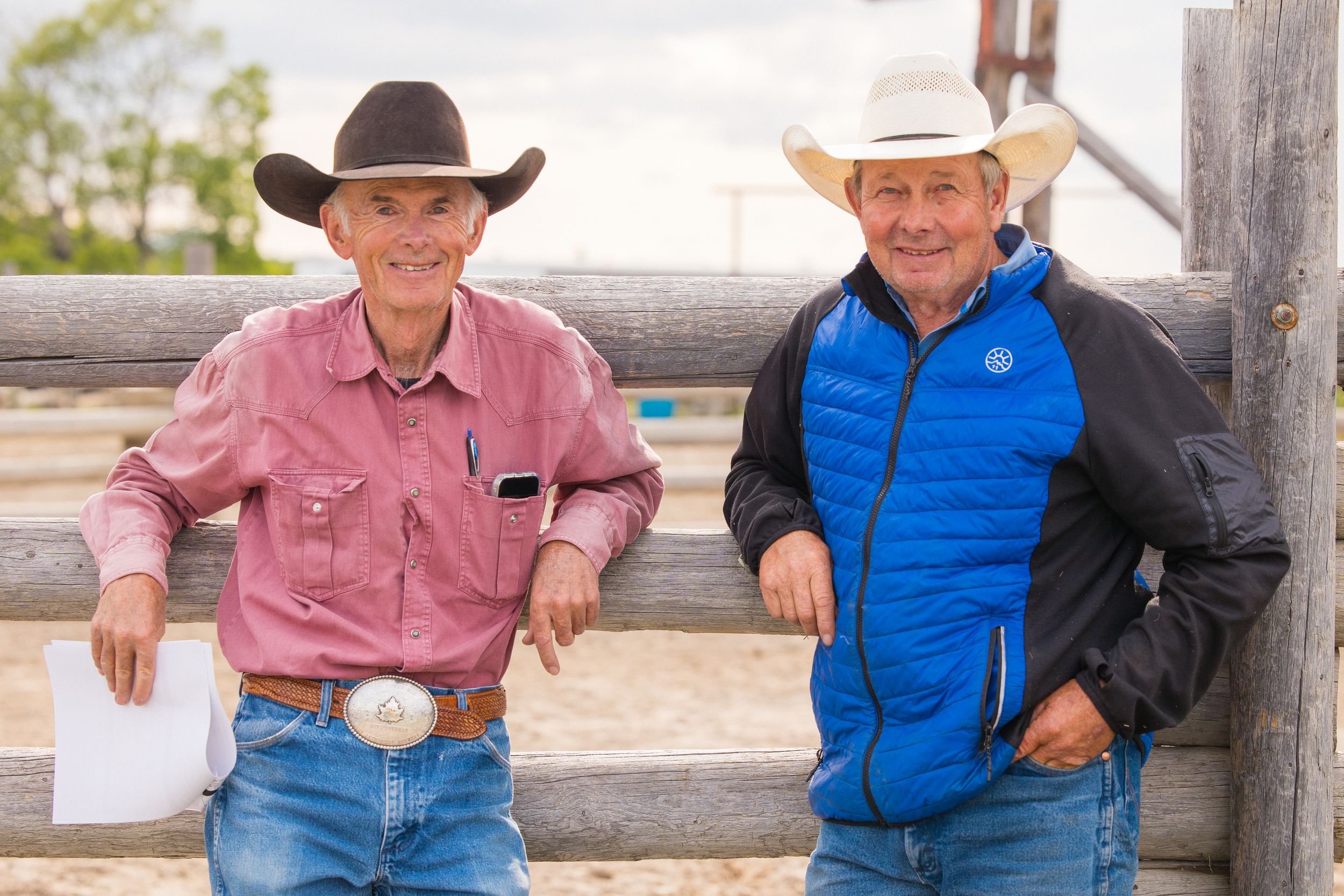Two smiling men in cowboy hats leaning on wooden ranch fence in sunny outdoor setting.
