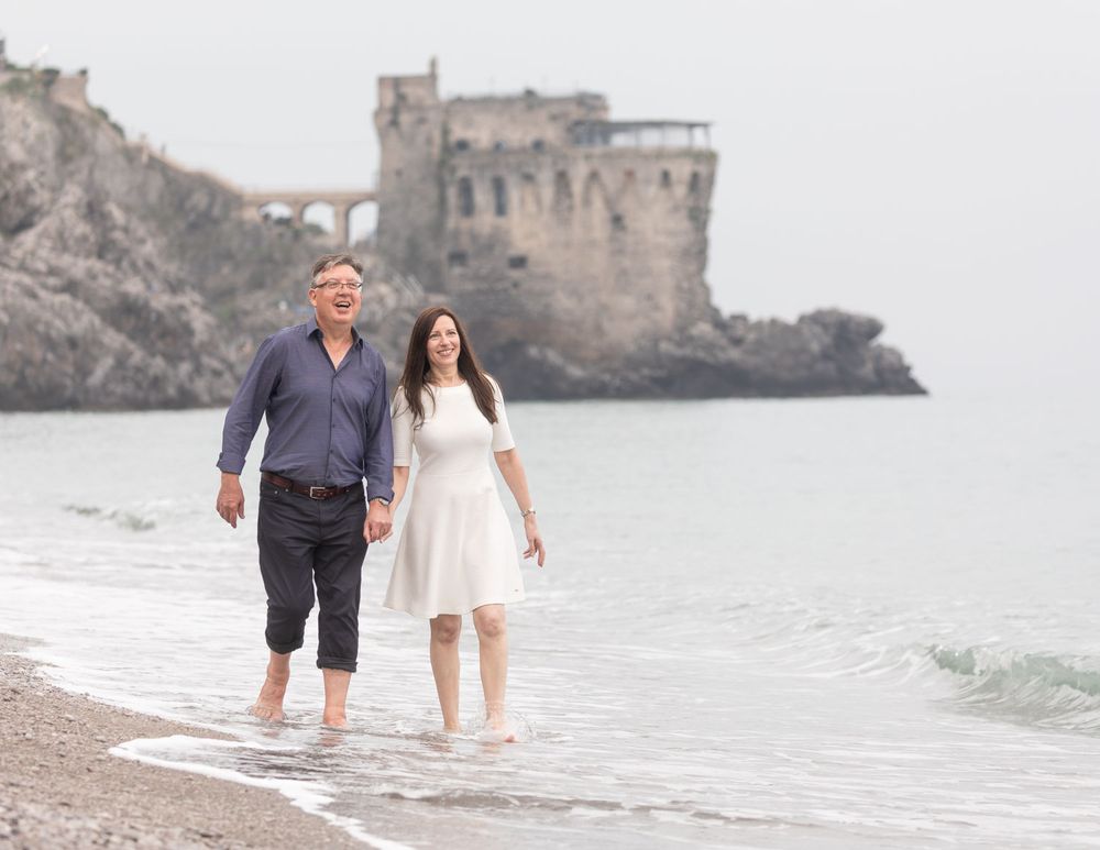 Couple walk along beach paddling in sea with historic castle in background