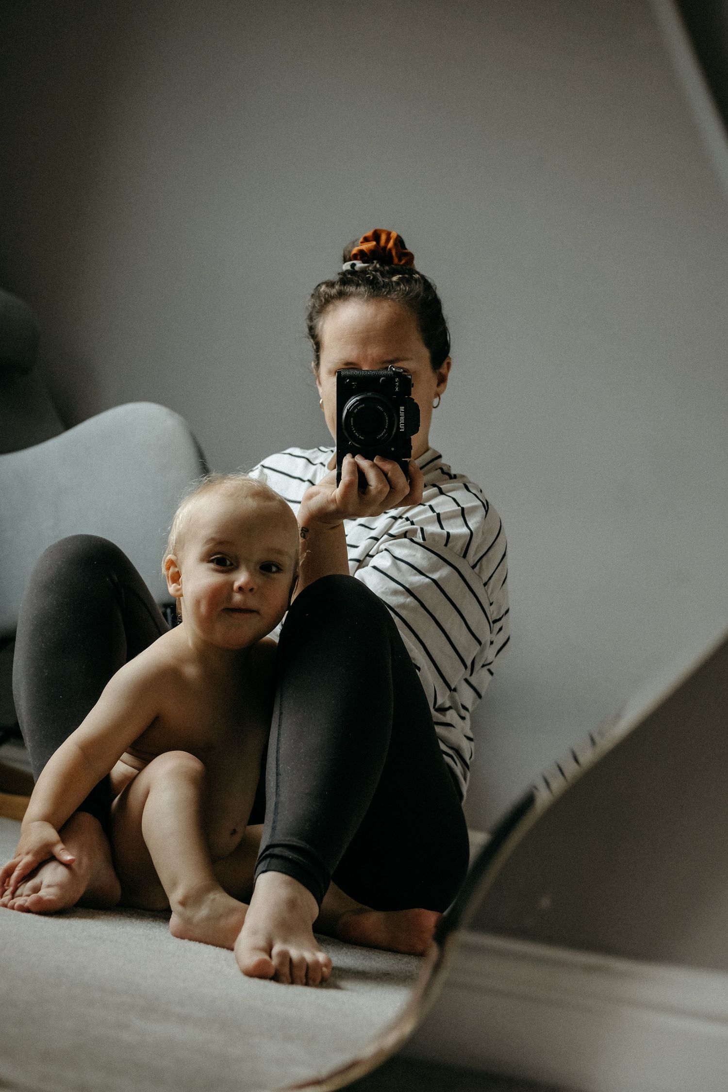 A mother seated on the floor smiling down at her baby during a relaxed, natural lifestyle newborn photography session.