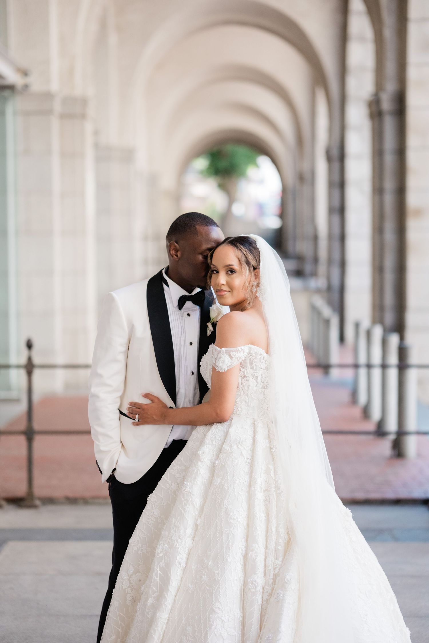 Elegant interracial couple posing under arched colonnade, bride in off-shoulder ballgown with veil, groom in white tuxedo.