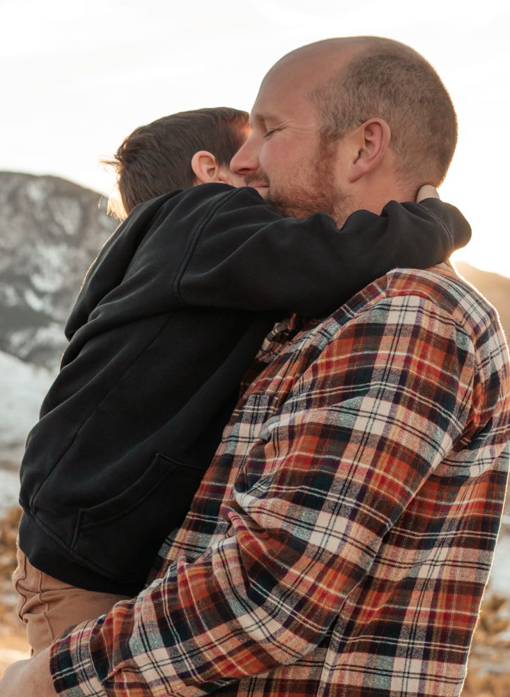 A bald man in a plaid shirt tenderly embraces a young child in a black hoodie outdoors | How I Help You Prep for Family Photos