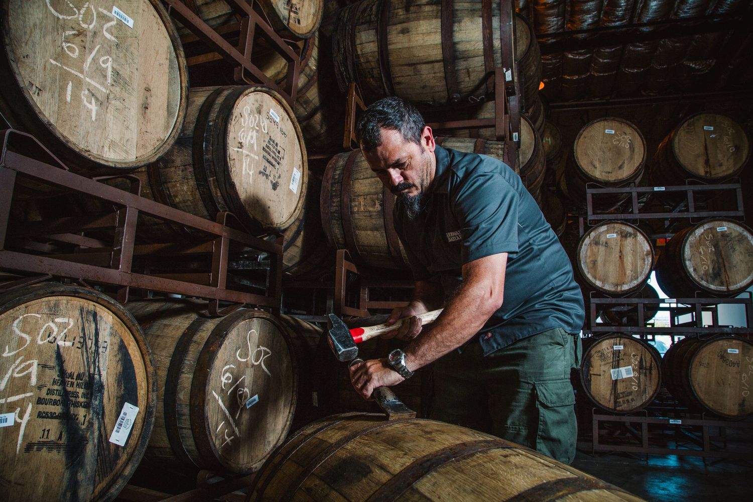 Worker in denim checking wooden whiskey barrels stacked in a traditional distillery warehouse.