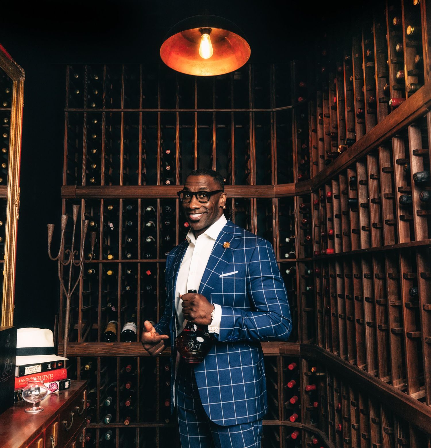 Person in blue checkered suit stands in elegant wine cellar with bottle racks and pendant lighting.
