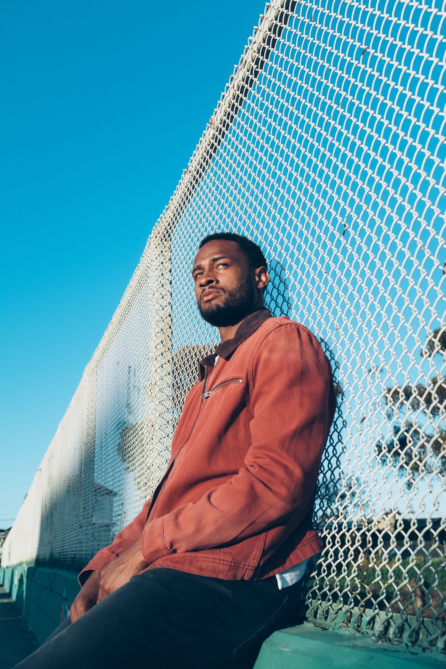 Person in an orange jacket stands against a chain link fence with bright blue sky in the background.