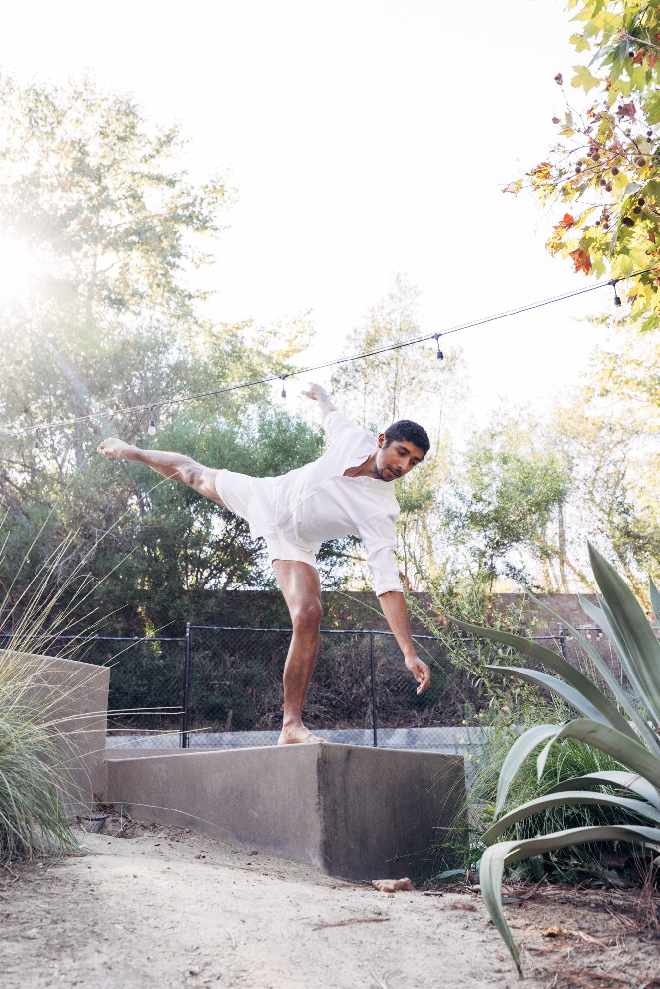 A dancer in white performs a graceful balancing pose outdoors in a sunlit garden setting.