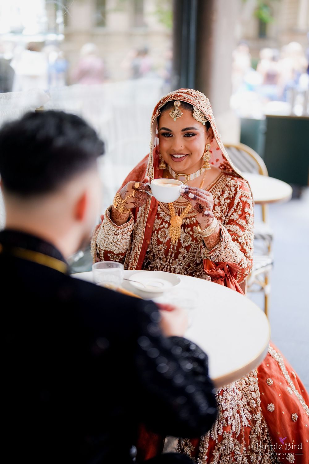 A couple shares tea at a cafe, with one person wearing an ornate red and gold traditional South Asian bridal outfit.