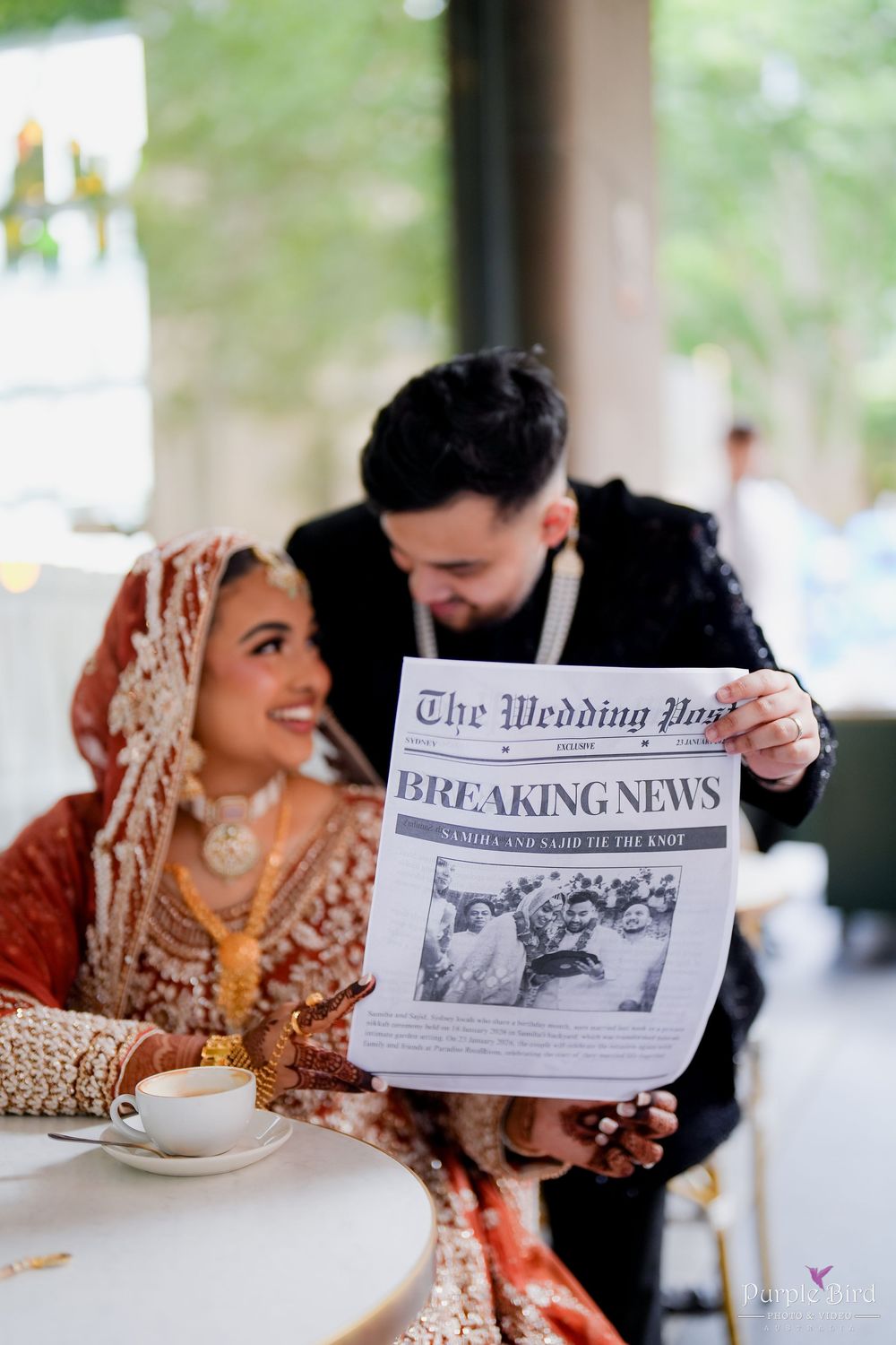 Couple reading custom wedding newspaper at reception table with coffee cups and traditional Indian bridal attire.