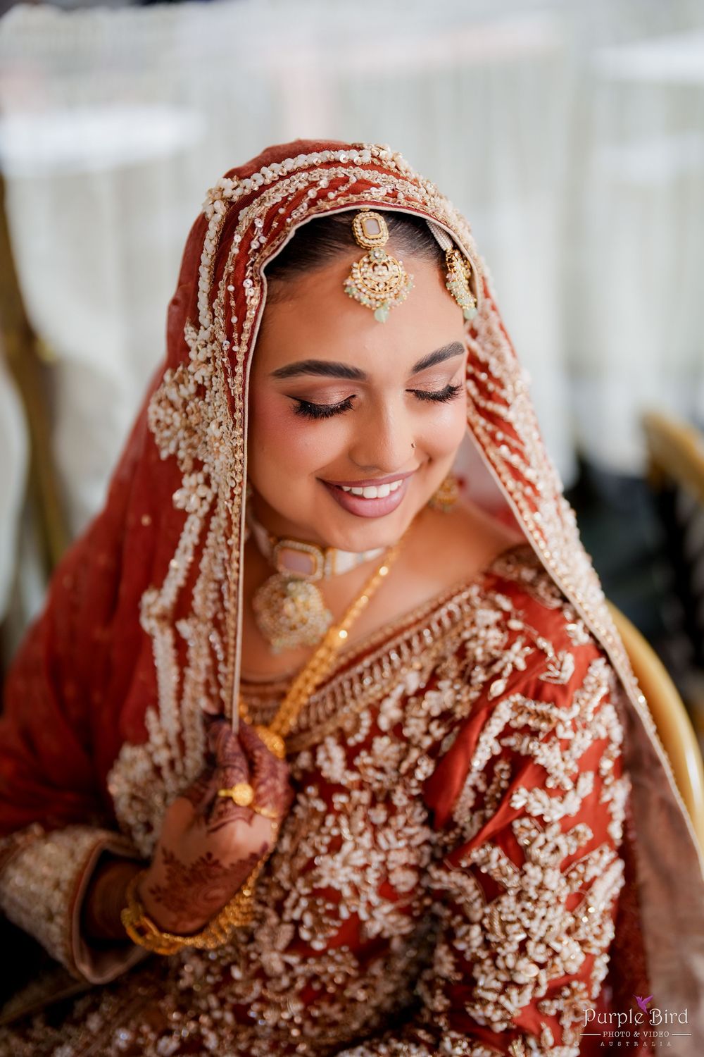 Traditional Indian bride in red lehenga with intricate gold embroidery and jewelry smiles during wedding ceremony.