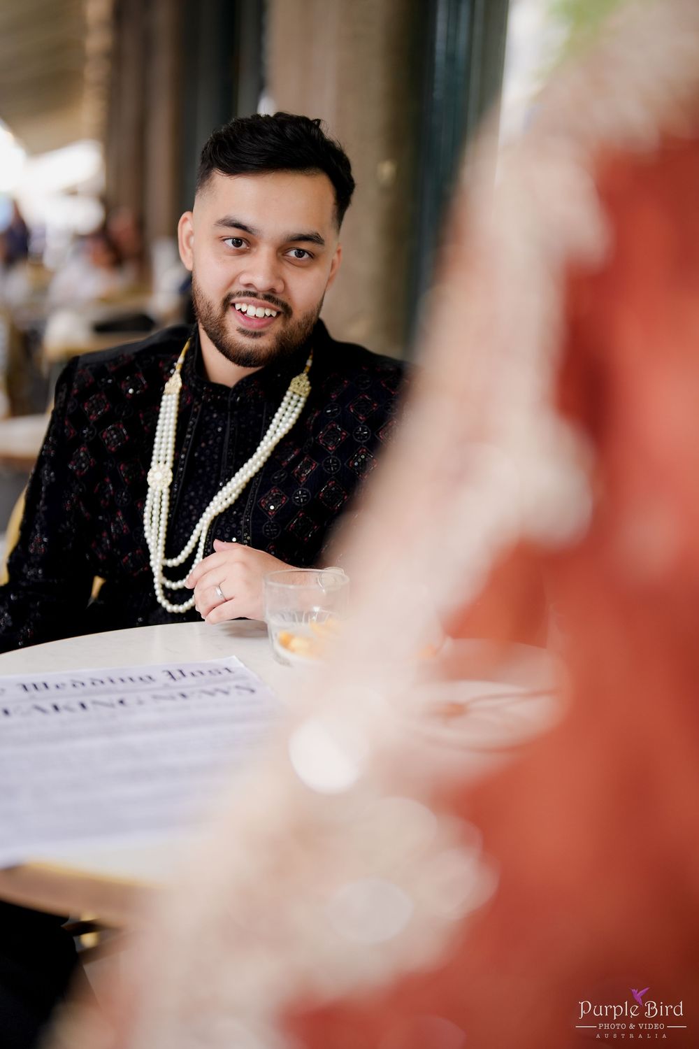A person in a black suit with pearl necklace signs documents at a formal event with soft focus foreground elements.
