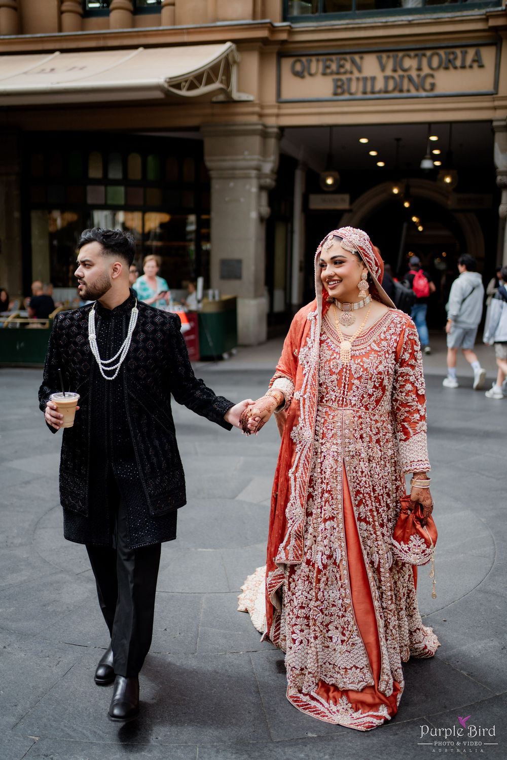 A couple in traditional wedding attire walks outside Queen Victoria Building, with the bride wearing a red embroidered dress.