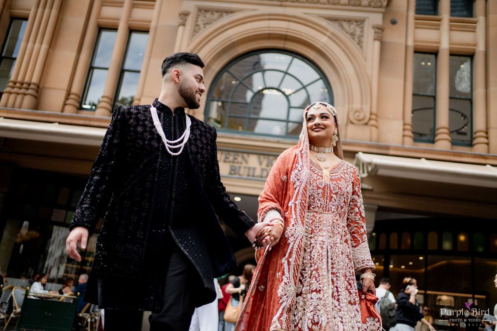 A couple in traditional attire poses for wedding photos in front of a grand historic building with arched architecture.