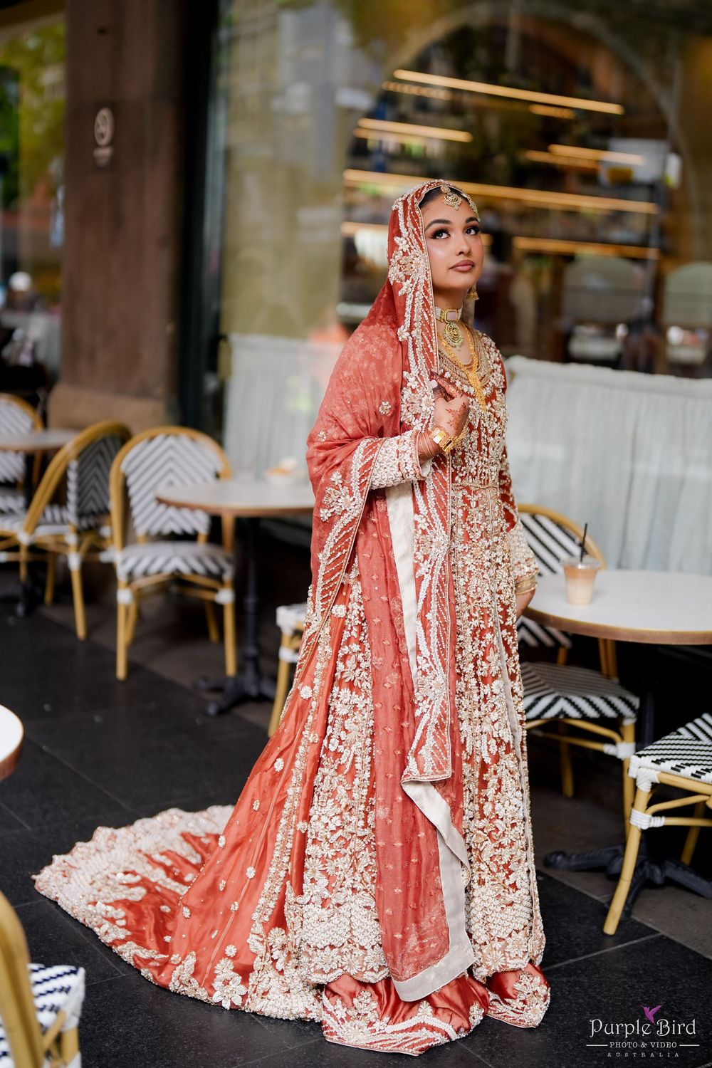 Traditional bride in coral pink and gold embroidered wedding attire sits elegantly in outdoor cafe setting.