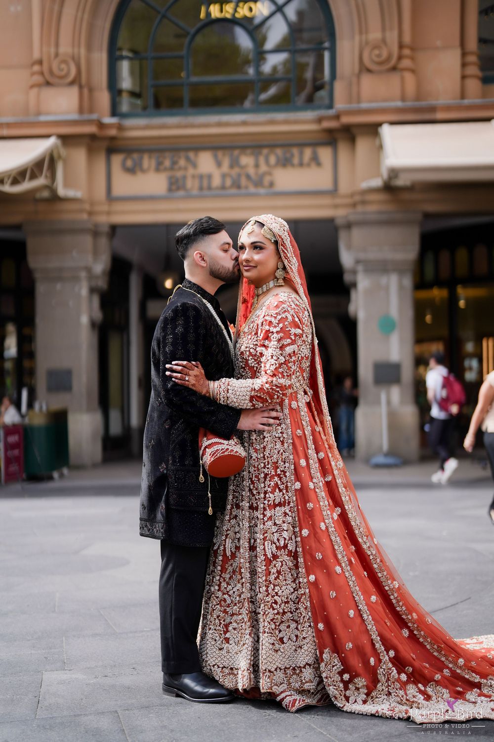 A couple in traditional South Asian wedding attire poses together outside a historic building.
