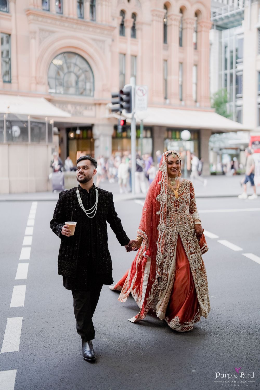 A couple in traditional South Asian wedding attire walks together on a city street near historic architecture.