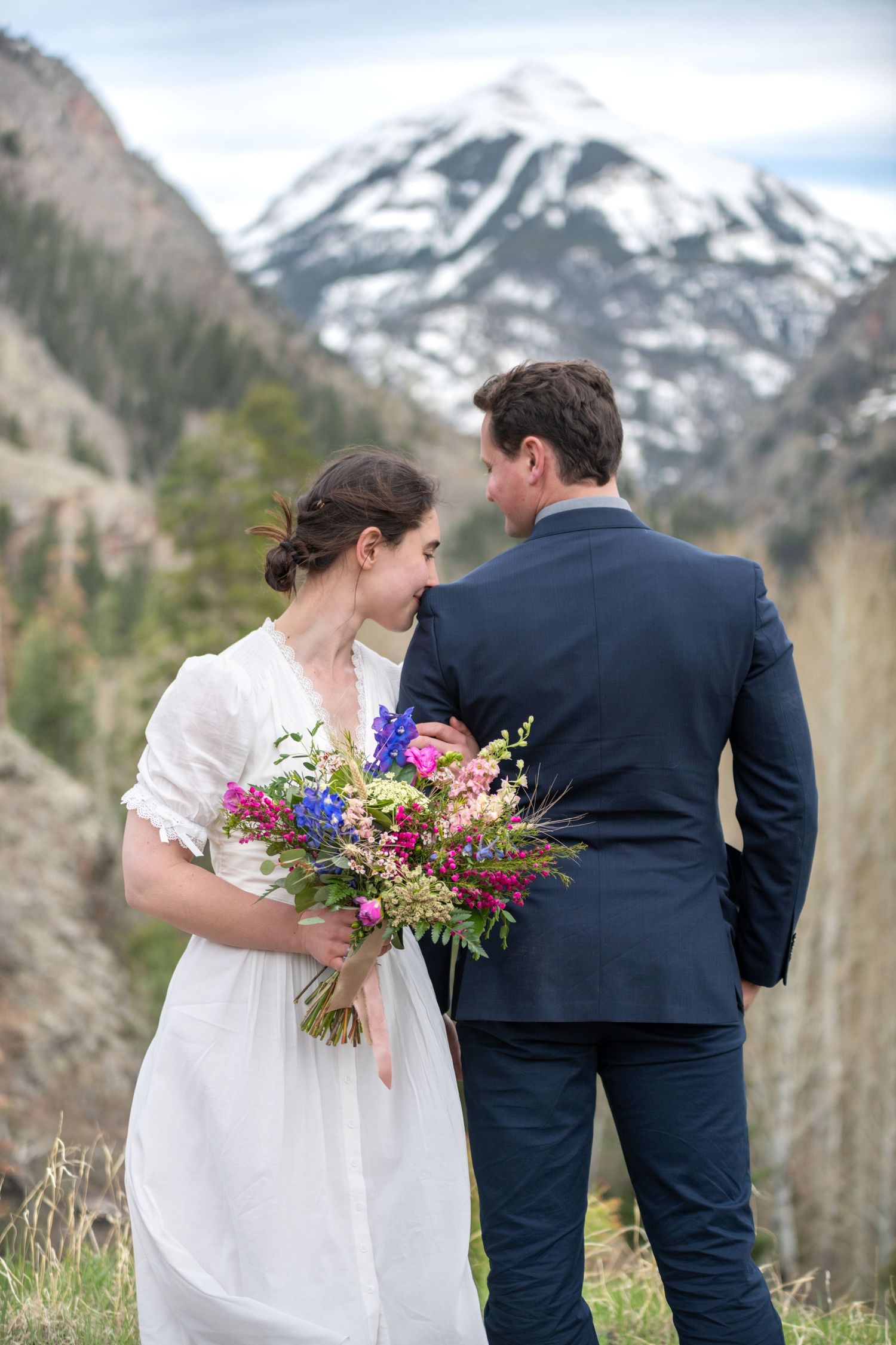 A bride is hugging the arm of her groom with a snowcapped mountain behind them near Ouray.  He is facing backward, she is facing forward.