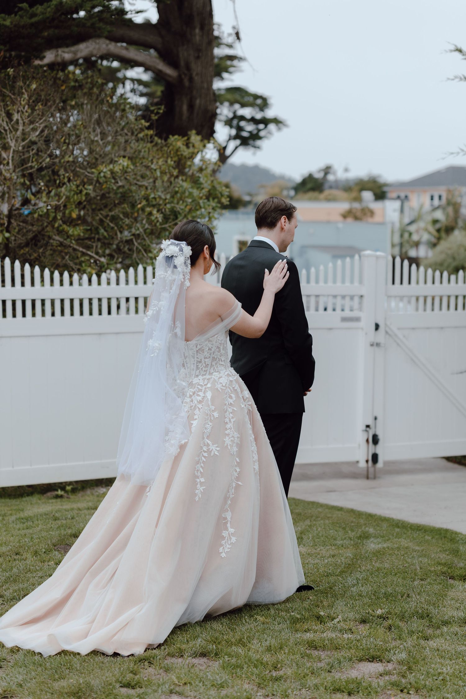 A couple in wedding attire stands together on a grassy lawn next to a white picket fence during their outdoor ceremony.