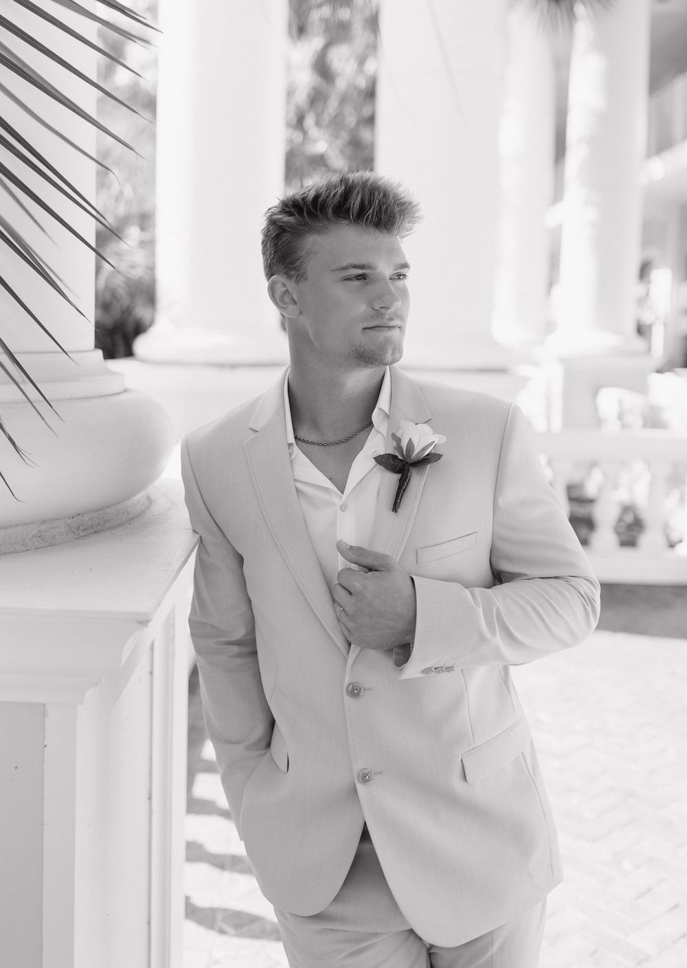 A groom in a light beige suit with a boutonniere poses elegantly against white columns in a black and white photo.