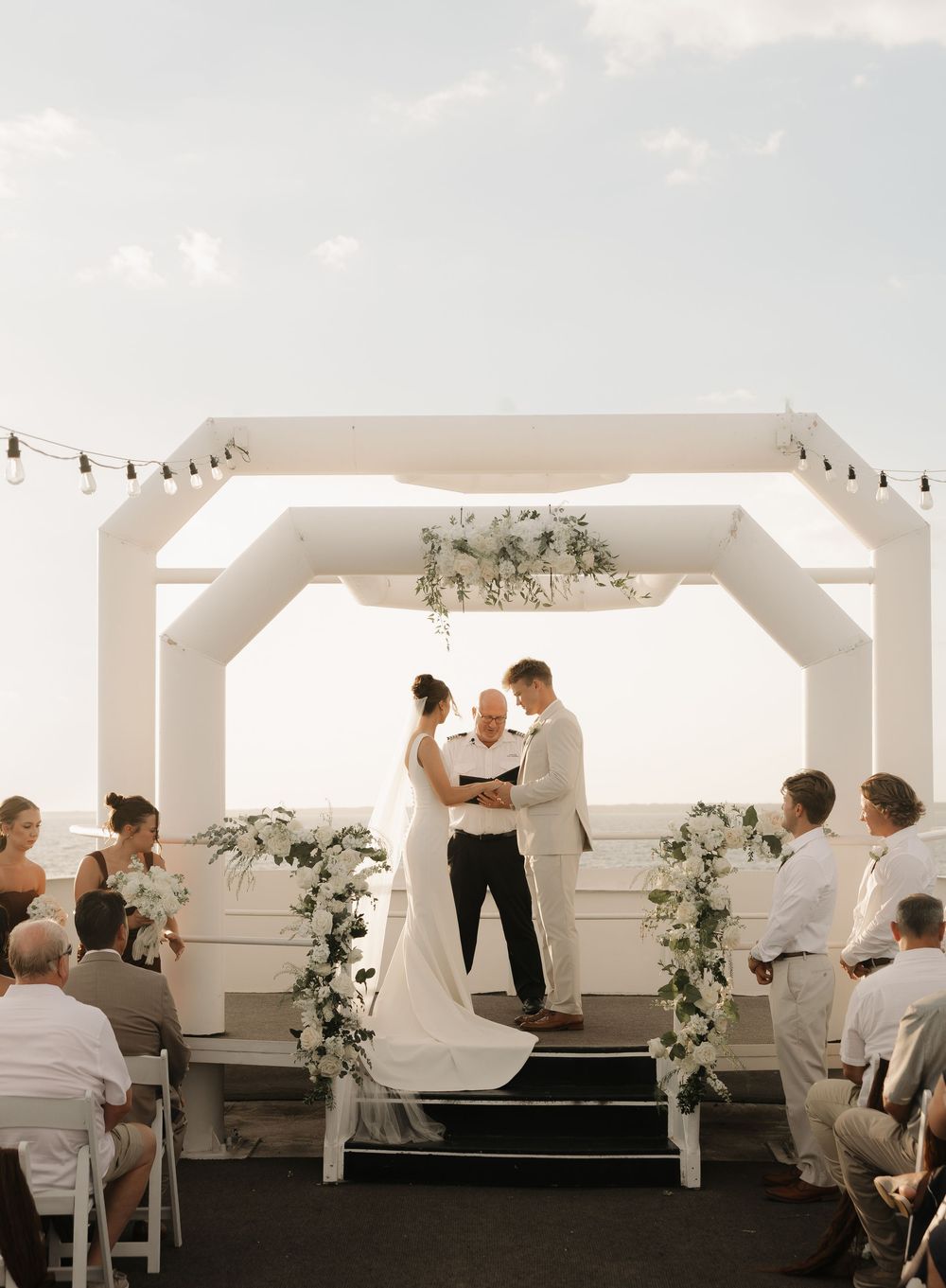 Elegant outdoor wedding ceremony under a white archway decorated with floral arrangements and greenery at sunset.