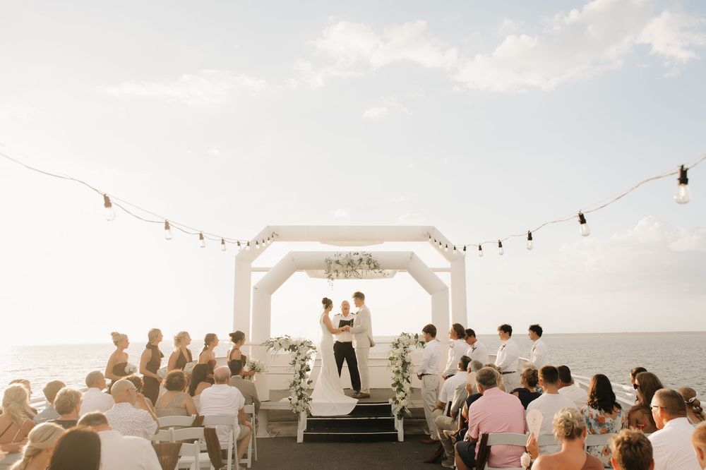 Wedding ceremony guests seated outdoors under string lights at sunset with an ocean view and white arch backdrop.