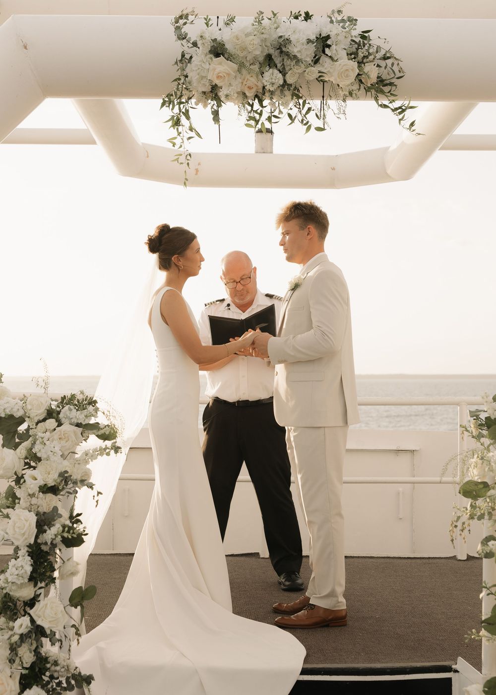 A wedding ceremony scene with floral decorations and white decor in a bright indoor venue.