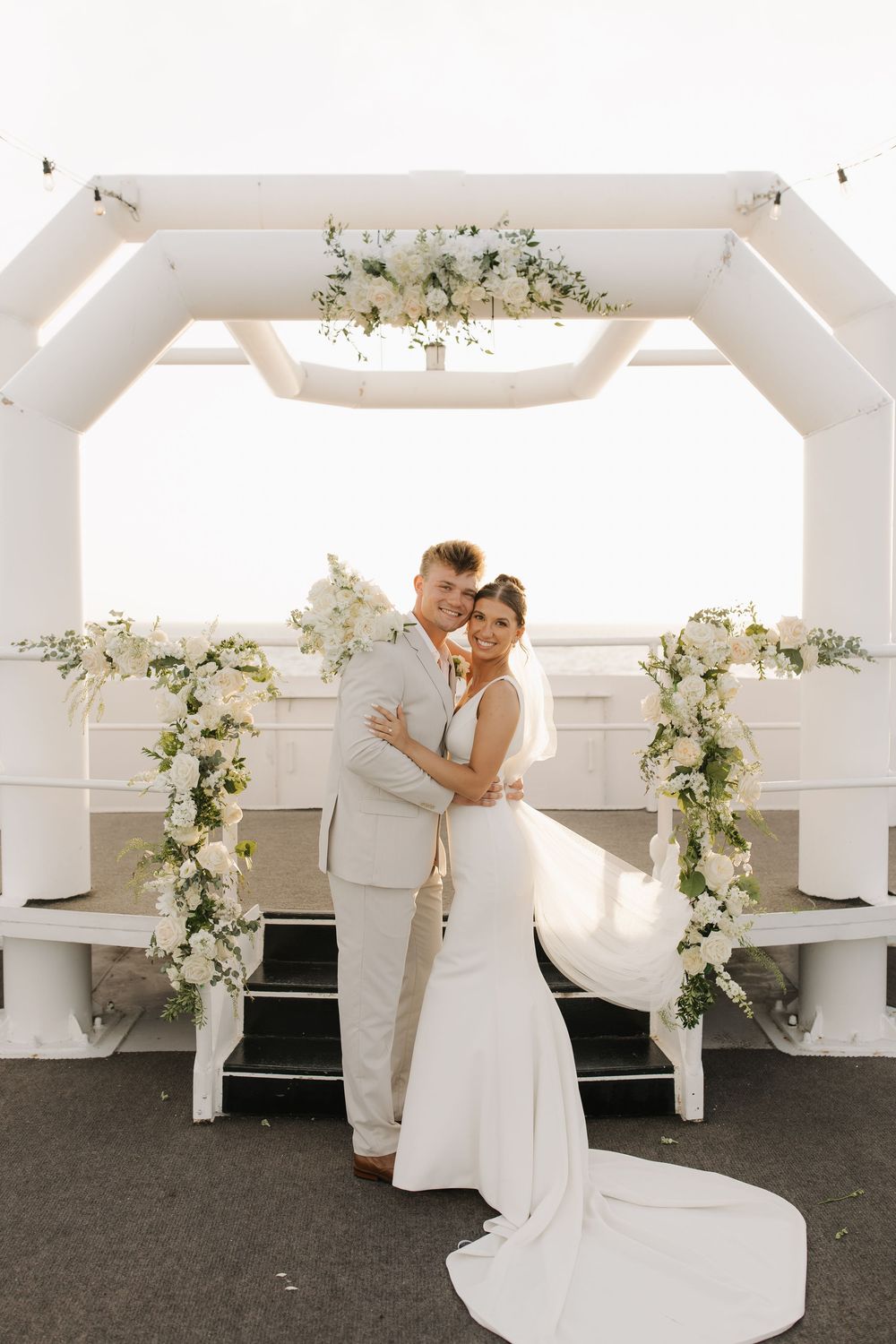 A couple in white dances in a bright indoor space decorated with floral arrangements.