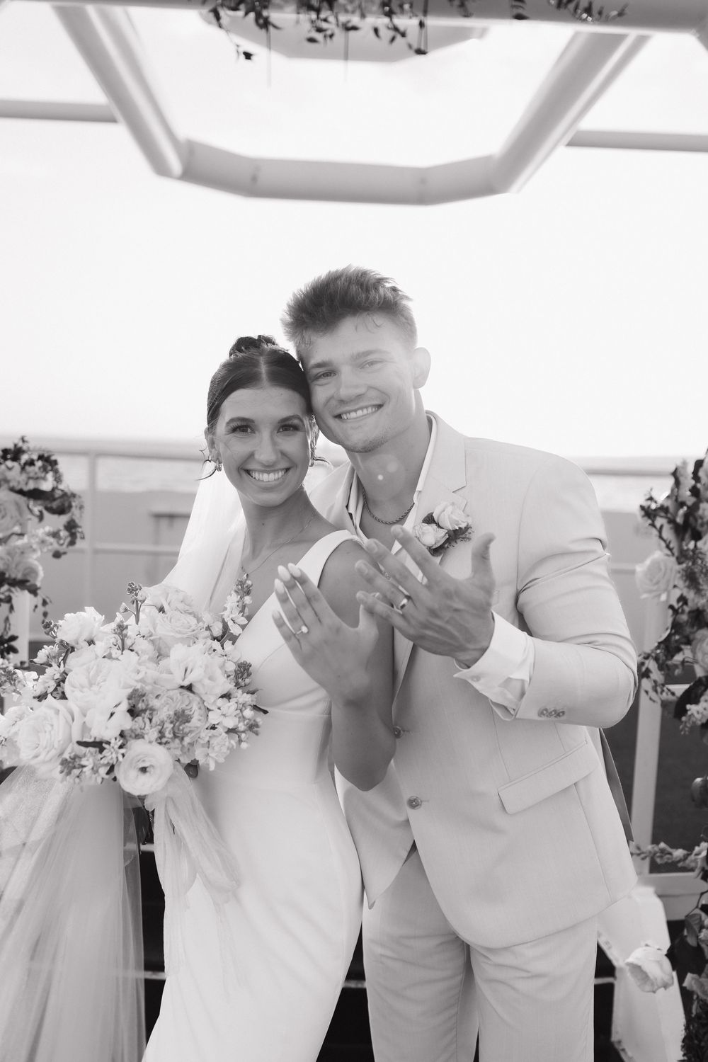 A newlywed couple shows off their rings while smiling under a floral-decorated wedding arch.