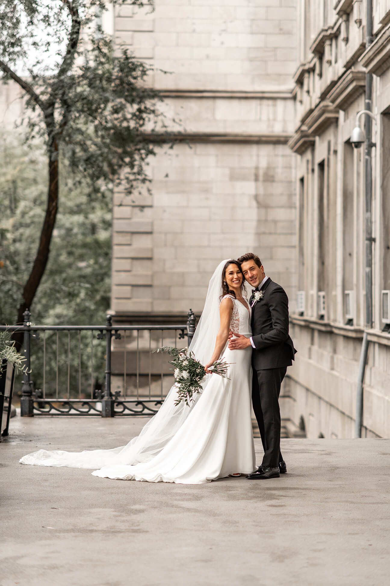 Elegant wedding couple embraces on stone balcony with classic architecture and greenery in background.  Montreal, Canada, Wedding Photograph