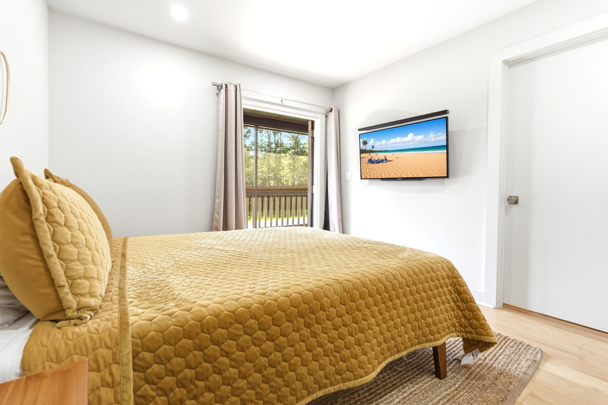 Cozy bedroom with golden yellow bedspread, wall-mounted TV, hardwood floors, and a window with outdoor views.