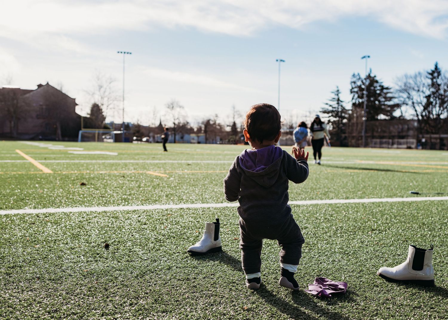A person crouches on an artificial turf sports field on a sunny day with stadium lights visible in the distance.