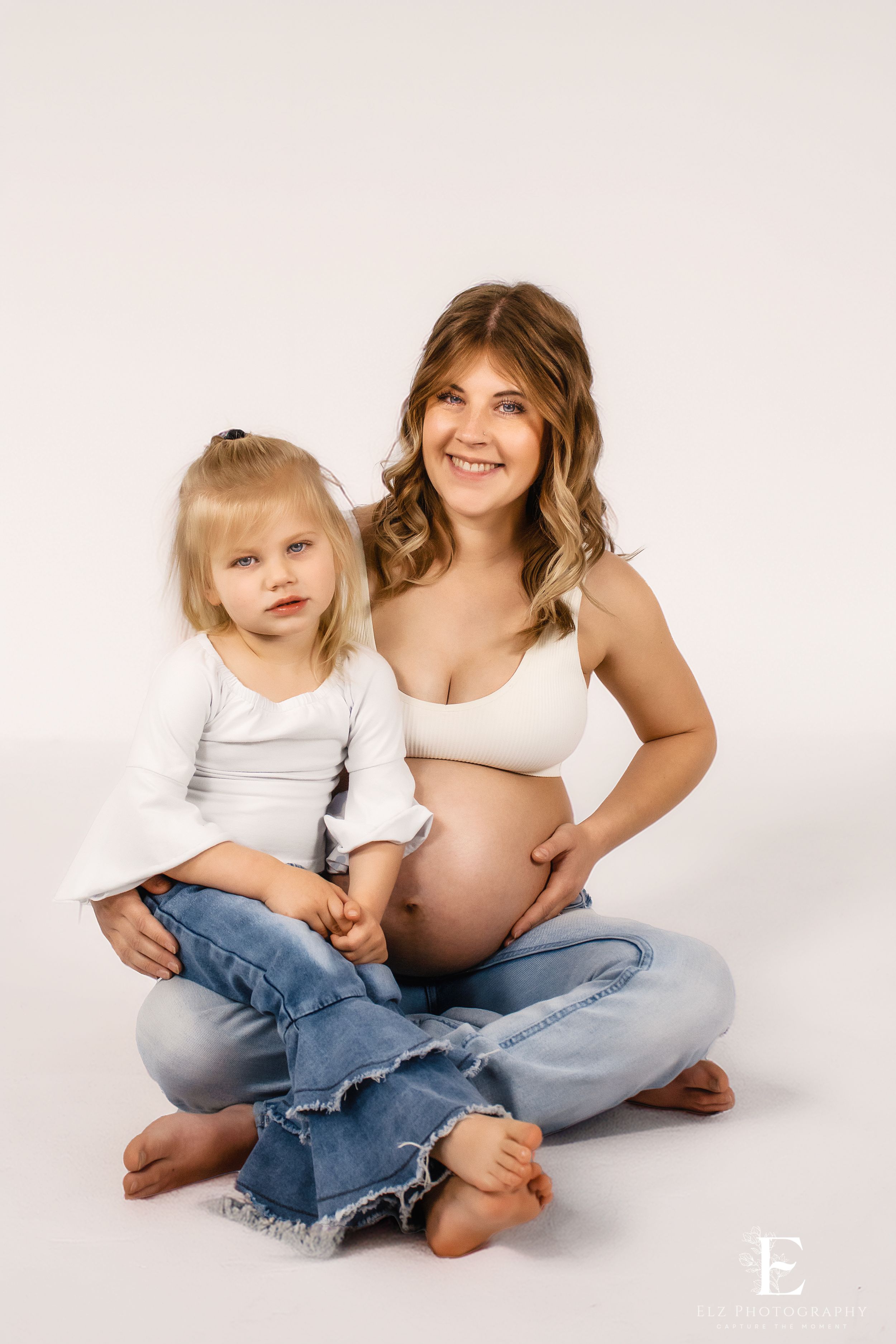 Maternity studio photos showing pregnant woman in white top and jeans sitting with small child on light background in Williston, ND.