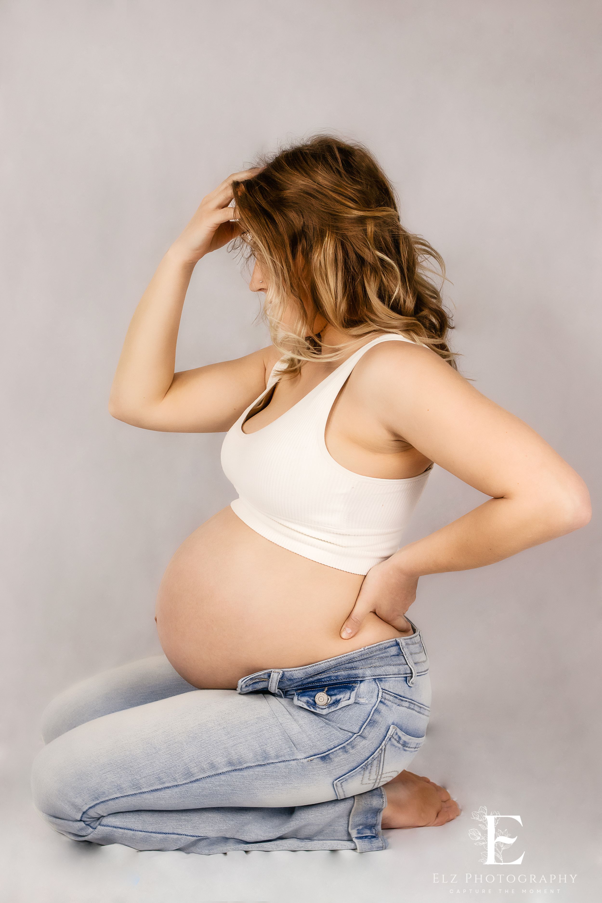 Artistic maternity photos showing a pregnant woman in jeans and white tank top against grey backdrop in Williston, North Dakota.