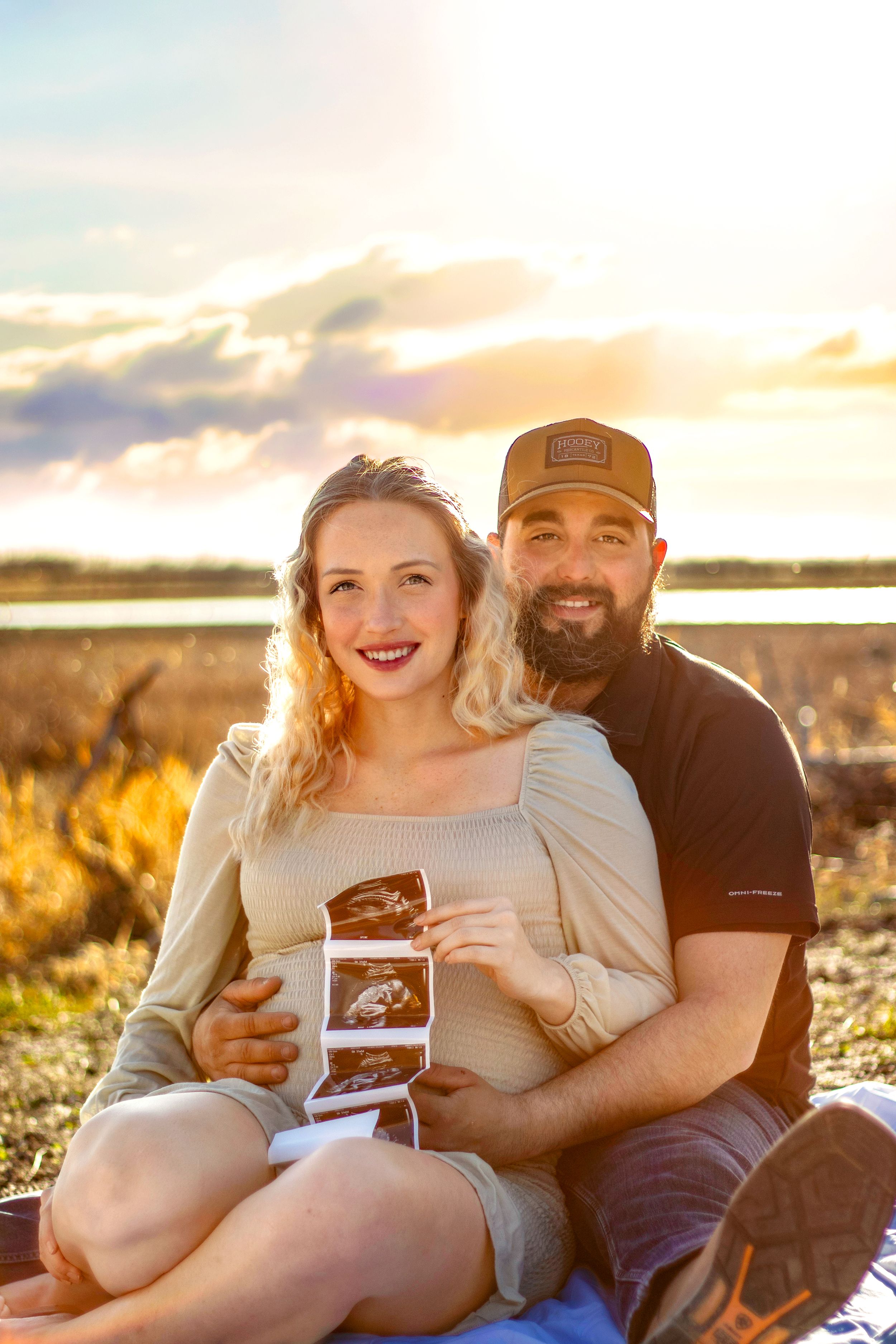 A couple sits together outdoors during golden hour sunset while holding ultrasound photos in Williston, North Dakota.