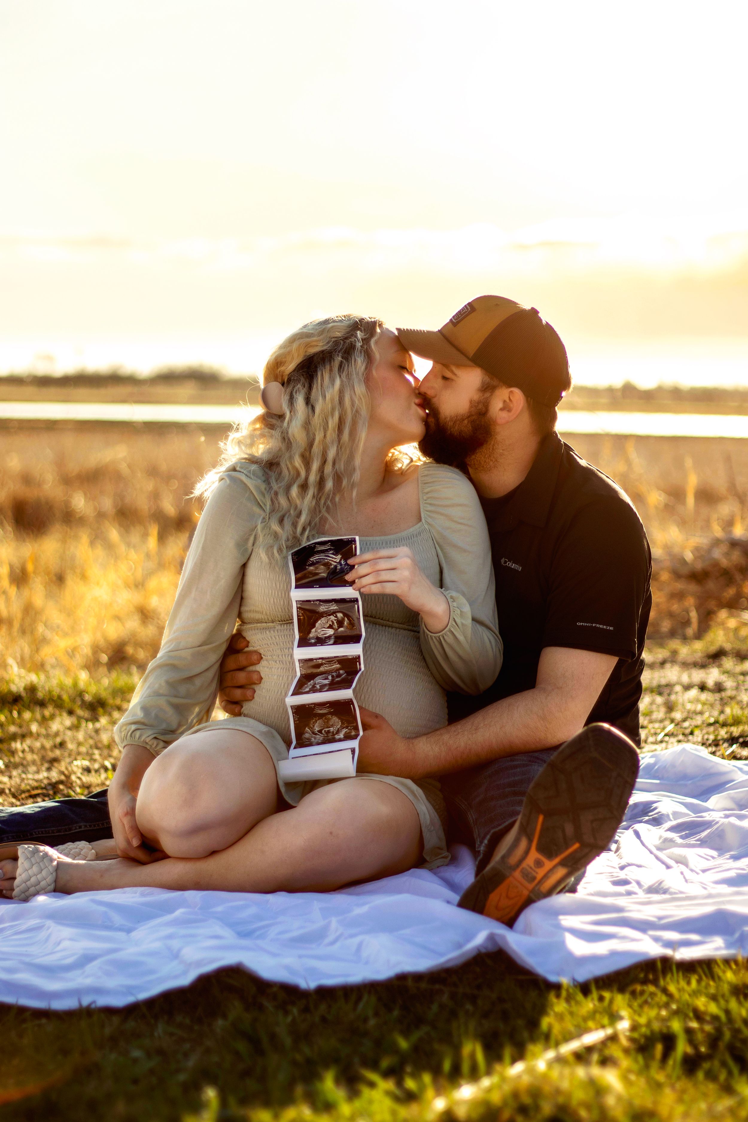 A couple sits on a blanket at sunset holding ultrasound photos during a pregnancy announcement photoshoot in Williston, North Dakota.