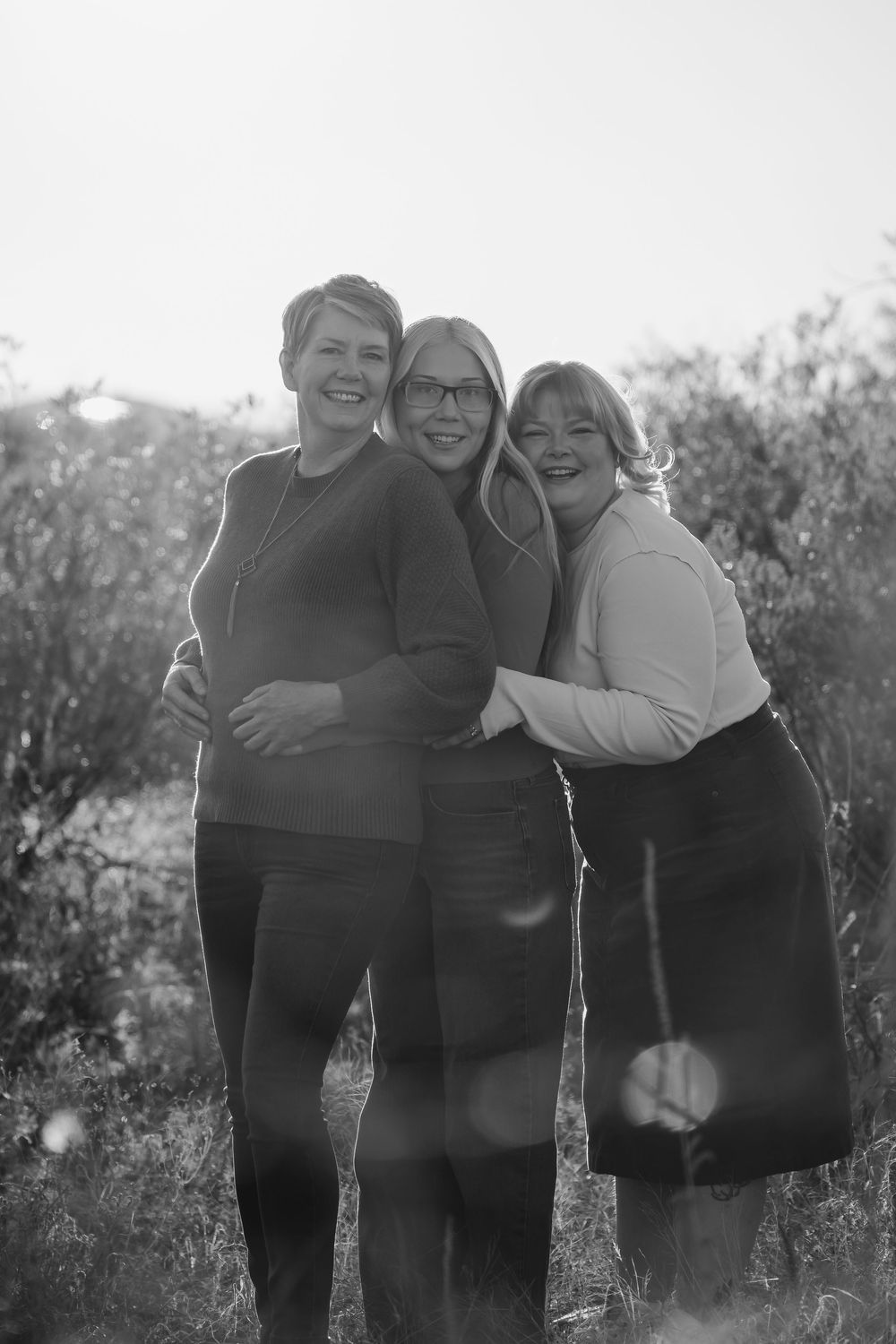A black and white portrait of three people embracing outdoors in a natural setting with sunlight behind them.