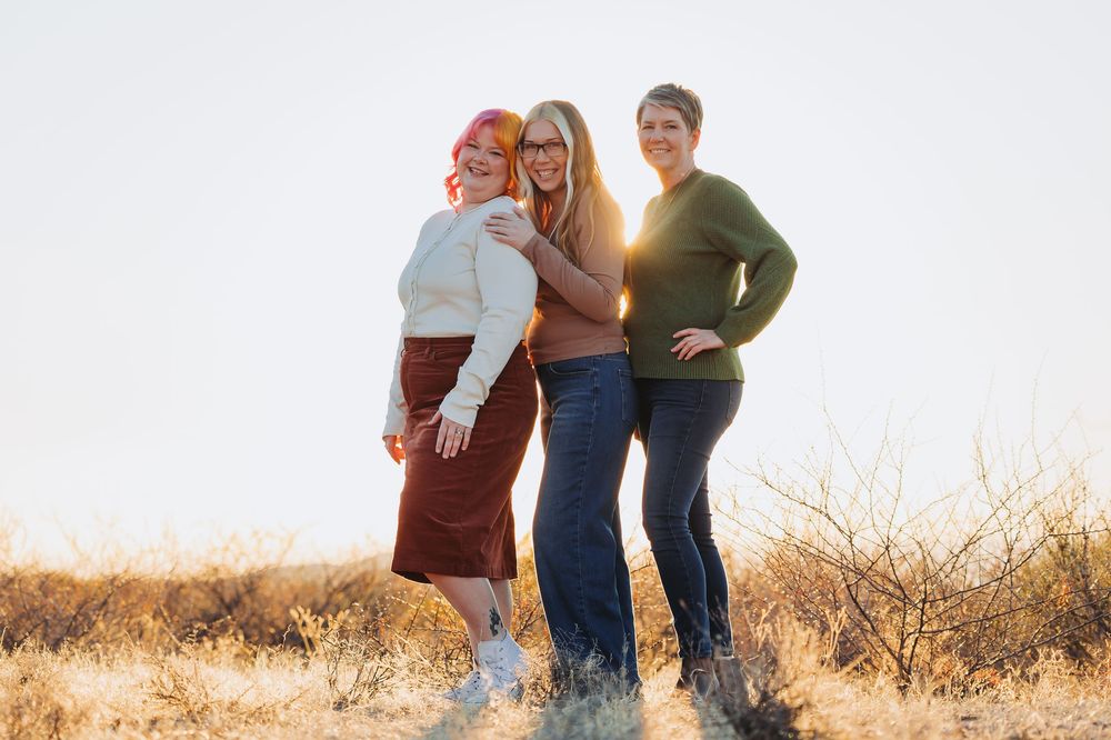 Three people stand together in a golden field at sunset creating silhouettes against the warm glowing sky.