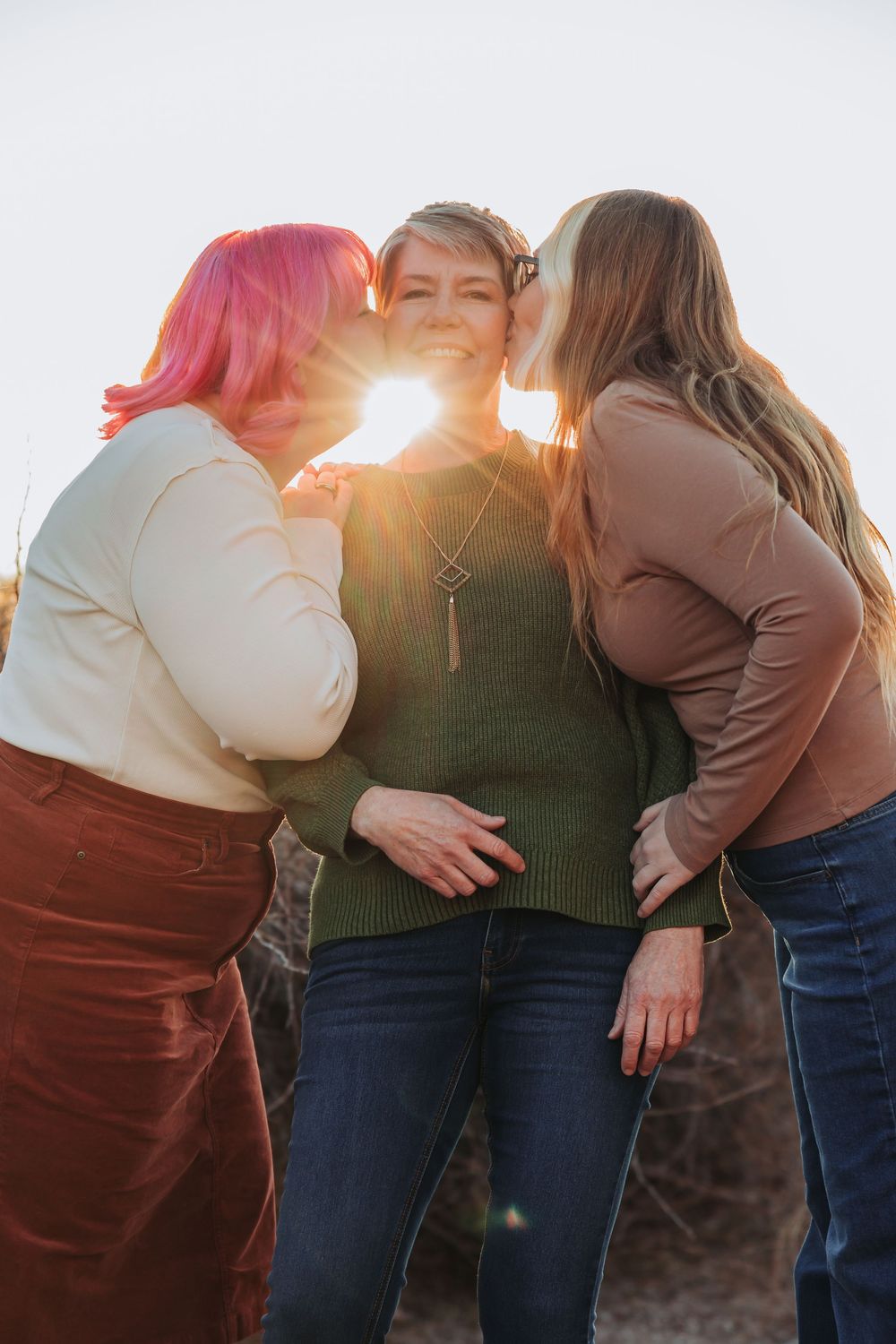 Three friends share an intimate moment at sunset with sunlight bursting between them against a bright sky.