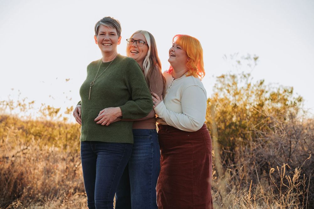 Three people embrace while standing in a golden field during sunset, creating a warm family portrait atmosphere.