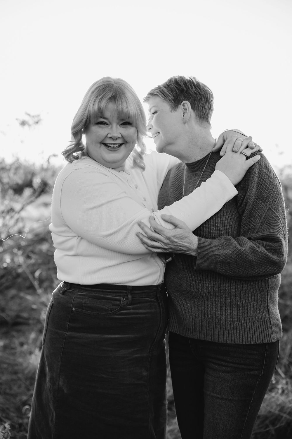 Black and white portrait of two people embracing and laughing together outdoors in casual sweaters.