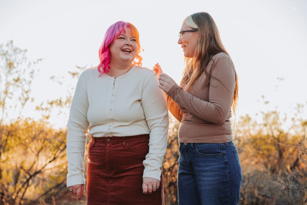 Two people laugh and smile together while wearing casual sweaters against a bright background.