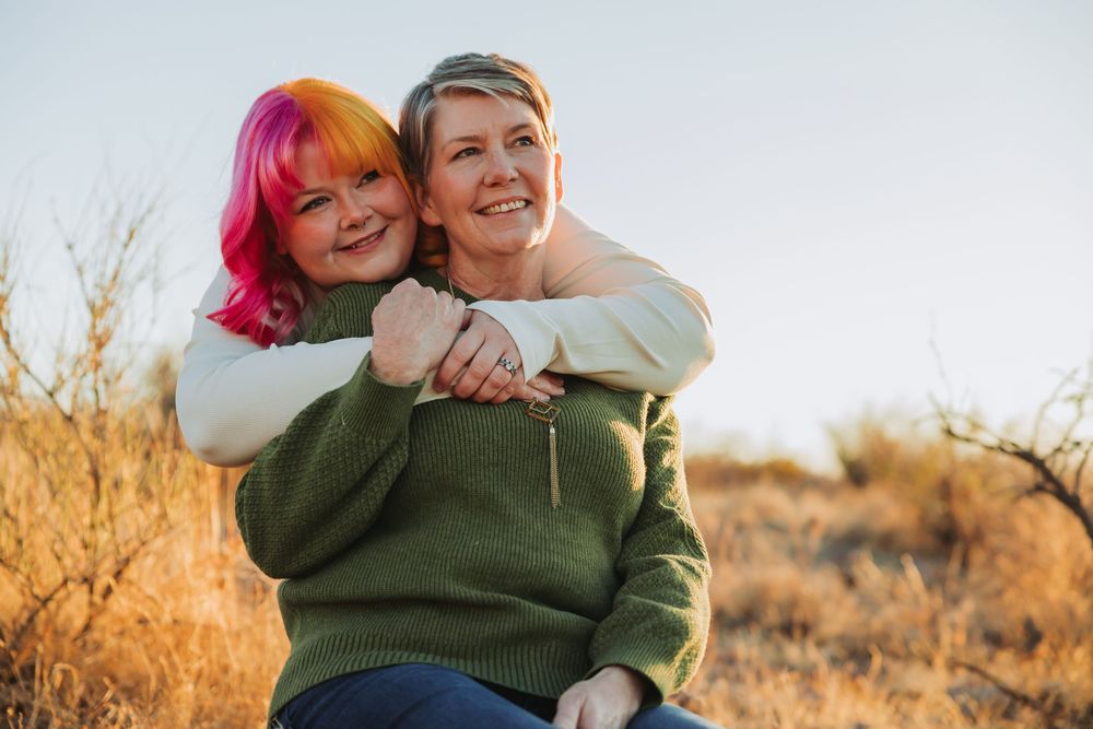 Three people share joyful hugs and laughter outdoors against a warm sunset backdrop.