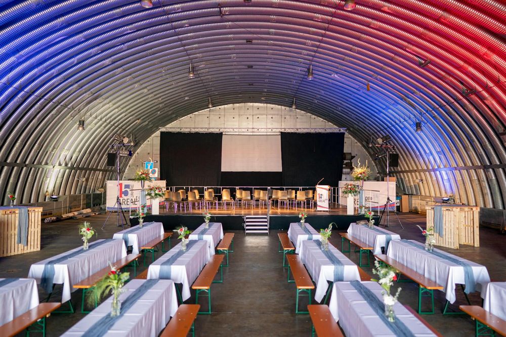 Long quonset hut interior with rows of wooden tables and benches illuminated by red and blue lighting.