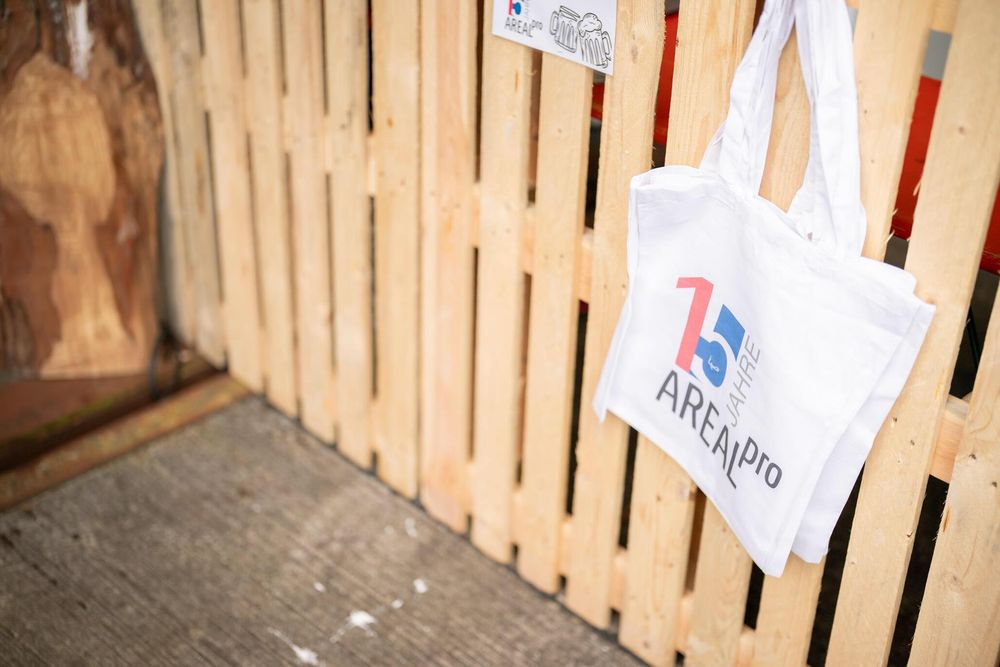 A white tote bag with a logo hangs on a wooden slatted fence or wall.