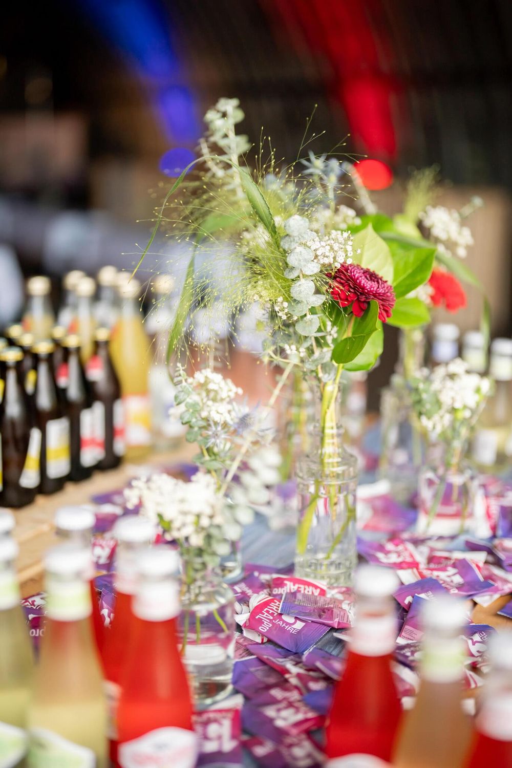 Glass vases with delicate floral arrangements and candles line a wedding reception table with wine bottles in the background.