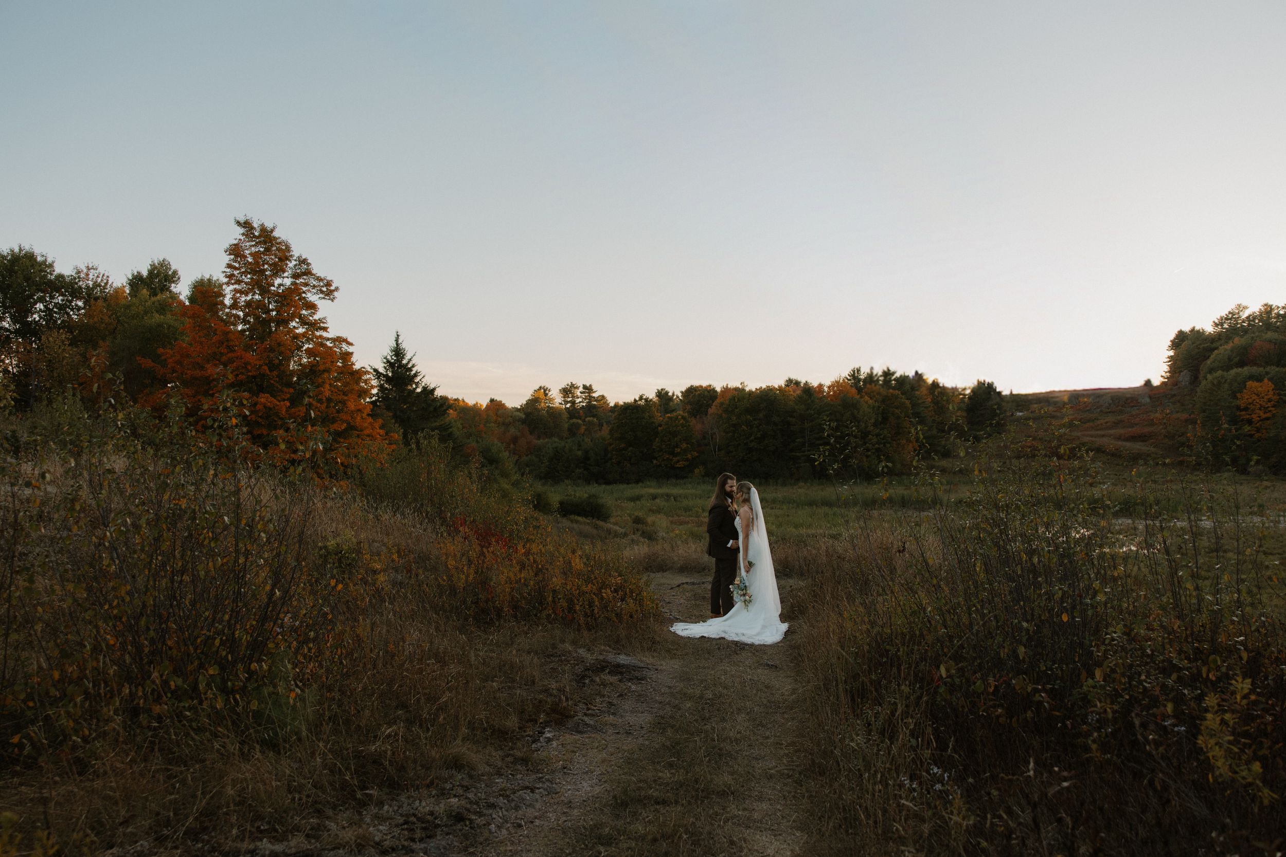 Wedding couple walks hand in hand through a scenic autumn field with golden trees at sunset.