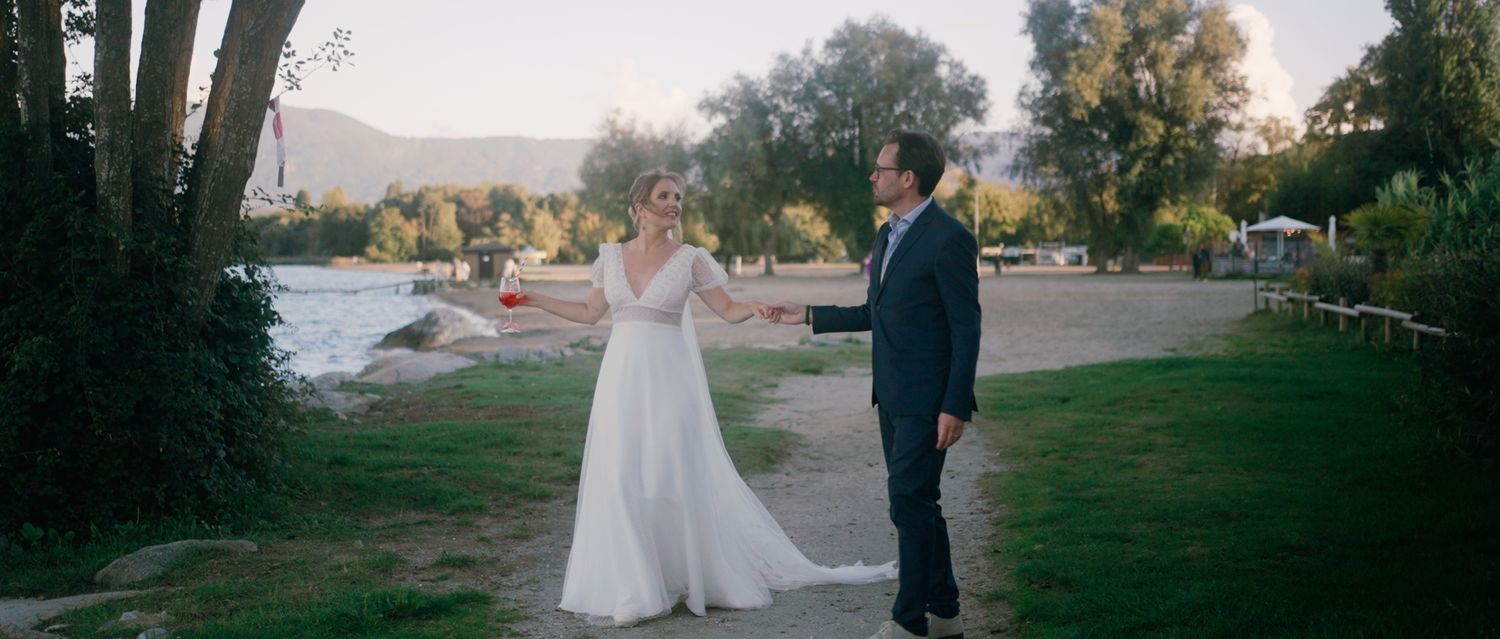 A couple in wedding attire stands together in a scenic outdoor setting with trees and natural lighting at sunset.